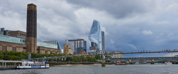 The image is an urban landscape photograph taken in London during a summer afternoon. The main subject is the Millennium Bridge spanning the River Thames, with pedestrians walking across its length. Prominently on the left stands the Tate Modern, a well-known contemporary art museum housed in a former power station and notable for its large brick chimney. The architecture of the scene includes modern high-rise buildings, among them One Blackfriars, also referred to as the Boomerang due to its distinctive curved design and reflective glass facade, which is visible in the centre background. The river Thames flows along the foreground, where a passenger boat is docked, emphasizing the busy ambience of this part of the city. The sky above is filled with dense clouds, indicative of typical summer weather in London. Overall, the photograph showcases key elements of London architecture and urban life along the Thames, with several iconic structures such as Tate Modern and One Blackfriars featured.